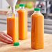 A hand pouring orange juice into 32 oz. tall square PET clear juice bottles with green lids.