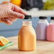 A hand holding a Square Milkman clear juice bottle with black lid filled with orange liquid.