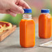 A hand holding a tall square clear PET juice bottle with orange juice in it with a blue lid.