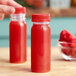 A hand holding a 4 oz. round clear plastic bottle with red liquid and a red lid.