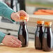 A person pouring liquid from a round PET clear carafe juice bottle with an orange lid.