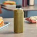 A 16 oz. tall square clear PET juice bottle filled with green liquid on a table next to a sandwich.