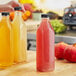 A hand pouring yellow juice from a 32 oz. clear square bottle with a black lid.