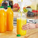 A hand pouring orange juice into a Square PET clear juice bottle with a green lid.