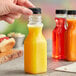 A hand holding a square clear PET juice bottle with orange juice inside and a black lid.