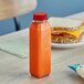 A 12 oz. clear rPET juice bottle with a red lid sits on a table next to a sandwich.