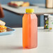 A 16 oz. tall square clear juice bottle with yellow liquid on a table next to a sandwich.