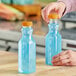 A woman's hand pouring blue liquid into a clear square PET juice bottle with an orange lid.