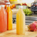 A person holding a glass of juice and a fruit next to a group of 32 oz. Tall Square Milkman PET Clear Juice Bottles.