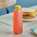 A sandwich and a bottle of orange juice sit on a table with a 12 oz. Tall Square rPET clear juice bottle with a yellow lid.
