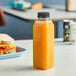 A 16 oz. tall square rPET clear juice bottle filled with orange juice on a table next to a sandwich.