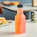 A close up of a 16 oz. square PET clear carafe juice bottle with a black lid on a table next to a sandwich.