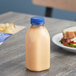 A 12 oz. clear square juice bottle with a blue lid next to a sandwich.
