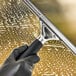 A gloved hand using an Ettore Master stainless steel squeegee with a black rubber grip to clean a window.