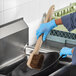 A person in blue gloves using a Lavex Palmyra bristled wok brush with a wood handle to clean a pan.