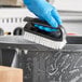 A hand in a blue glove using a Lavex black iron scrub brush to clean a black object.