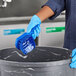 A person in blue gloves holding a Lavex blue utility brush over a bucket.