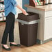 A woman standing next to a Lavex brown square trash can with a plastic bag over it.