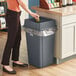 A woman standing in front of a Lavex gray plastic trash can with a plastic bag over it.