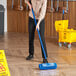 A woman using a Lavex blue bi-level floor scrub brush with a blue metal handle to scrub a floor.