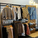A UDIZINE black wall-mount retail clothing display with faceouts and hanging rails for shelves holding clothes.