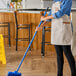 A woman in an apron using a blue Lavex metal threaded broom handle to clean a floor in a professional kitchen.