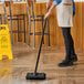 A person using a Lavex black deck brush with a squeegee to clean a floor.