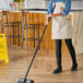 A woman in a white apron sweeping the floor with a Lavex black metal broom handle.
