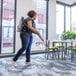 A woman uses a ProTeam cordless backpack vacuum to clean the floor in a room with tables and chairs.