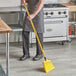 A man in an apron sweeping a floor with a yellow angled broom.