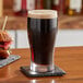 A glass of dark liquid on a coaster on a table with a sandwich.