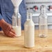 A person pouring white liquid into a 16 oz. clear PET sauce bottle with a blue cap.