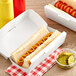 A hot dog in a white paper clamshell take-out box.