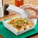 A hand holding a 6" x 6" white paper clamshell with nachos inside.