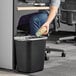 A person putting a paper cup into a Lavex rectangular black trash can in an office.