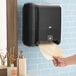 A hand holding a paper towel in a black Tork Elevation Matic automatic paper towel dispenser.