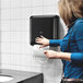 A black wall-mounted automatic hand towel roll dispenser being used in a restroom.