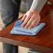 A person cleaning a table with a blue Advantage Supplies microfiber cloth.