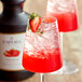 A glass of red liquid with a strawberry garnish next to a bottle of Capora Strawberry Flavoring Sauce.