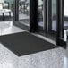 A black Lavex waffle entrance mat on a floor in front of a glass door.