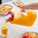 A hand pouring orange juice from a Rubbermaid Mixermate pitcher with a white lid into a glass.