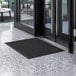 A black Lavex Needle Rib entrance mat on a white tiled floor in front of a glass door.