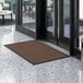 A brown Lavex Needle Rib entrance mat placed on a white floor in front of a glass door.
