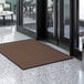 A brown Lavex Needle Rib entrance mat on a marble floor in front of a glass door.
