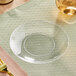 A clear Visions plastic coupe plate on a table with a glass of liquid.