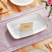 A Visions white plastic rectangular coupe platter with a gold band and a place card on it.