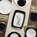 A white table set with a Visions white square plastic plate with a silver border and silverware.