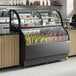 An Avantco black curved dual service refrigerated air curtain merchandiser with drinks and pastries on shelves.