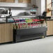 An Avantco black horizontal air curtain merchandiser on a hospital cafeteria counter with bottles of soda inside.
