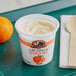 A white September Farm Peach yogurt cup on a table with an orange and a spoon.